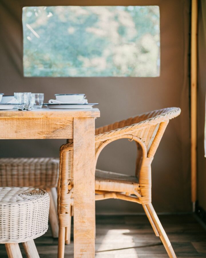 A rustic outdoor dining setup at Exe Valley Glamping features a wooden table and wicker chairs under a beige canopy. Sunlight filters through a clear plastic window, illuminating plates and glasses ready for a meal. In the background, blurred greenery completes the serene scene.