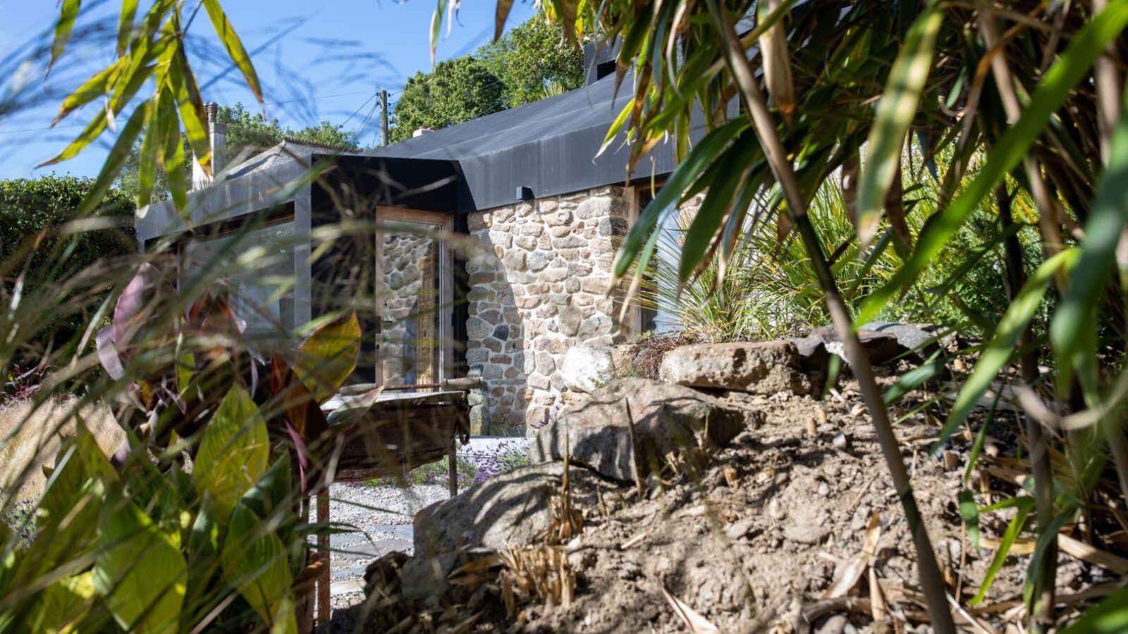 A stone house with large glass windows is partially obscured by lush greenery in the foreground. The roof appears to be metal, and there is a small, rocky area with plants in front. Sunlight filters through the trees, creating a serene, natural atmosphere reminiscent of a fallen angel's sanctuary.