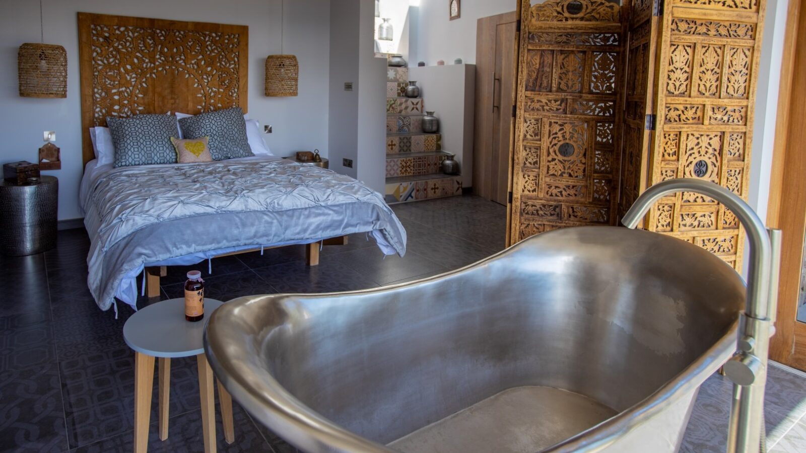 A cozy bedroom with a modern metal bathtub in the foreground exudes the charm of a fallen angel’s sanctuary. The room features a double bed with a carved wooden headboard, decorative wooden screens, and steps leading to an open area, merging rustic and contemporary elements seamlessly.