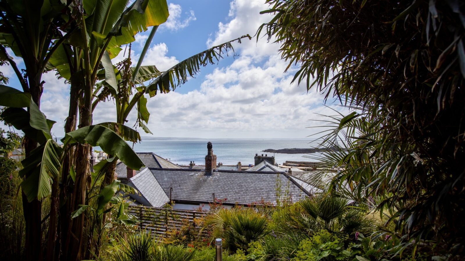 View of a coastal village with slate-roofed houses, seen through tropical greenery including banana trees. The background features a beach and a vast, cloudy sky over a calm sea. The scene feels like a hidden paradise touched by the brush of a fallen angel.