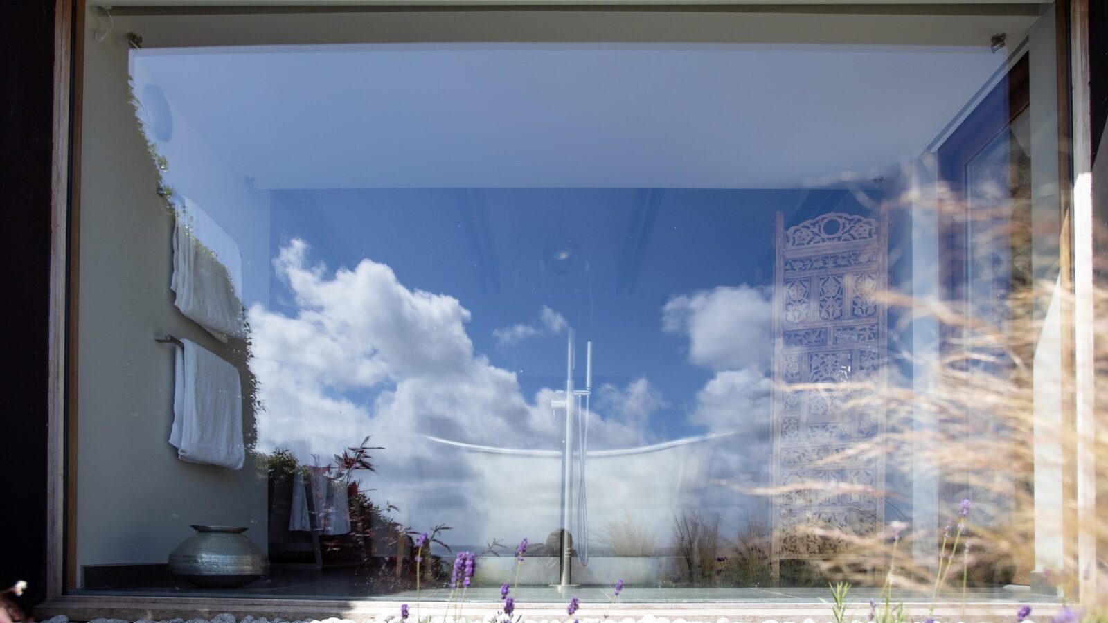 A large glass window reflects a clear blue sky with scattered white clouds. Inside, a modern white bathtub is visible along with a decorative screen and towels hanging on the wall. The foreground has a garden with small white pebbles, purple flowers, and an angel sculpture.