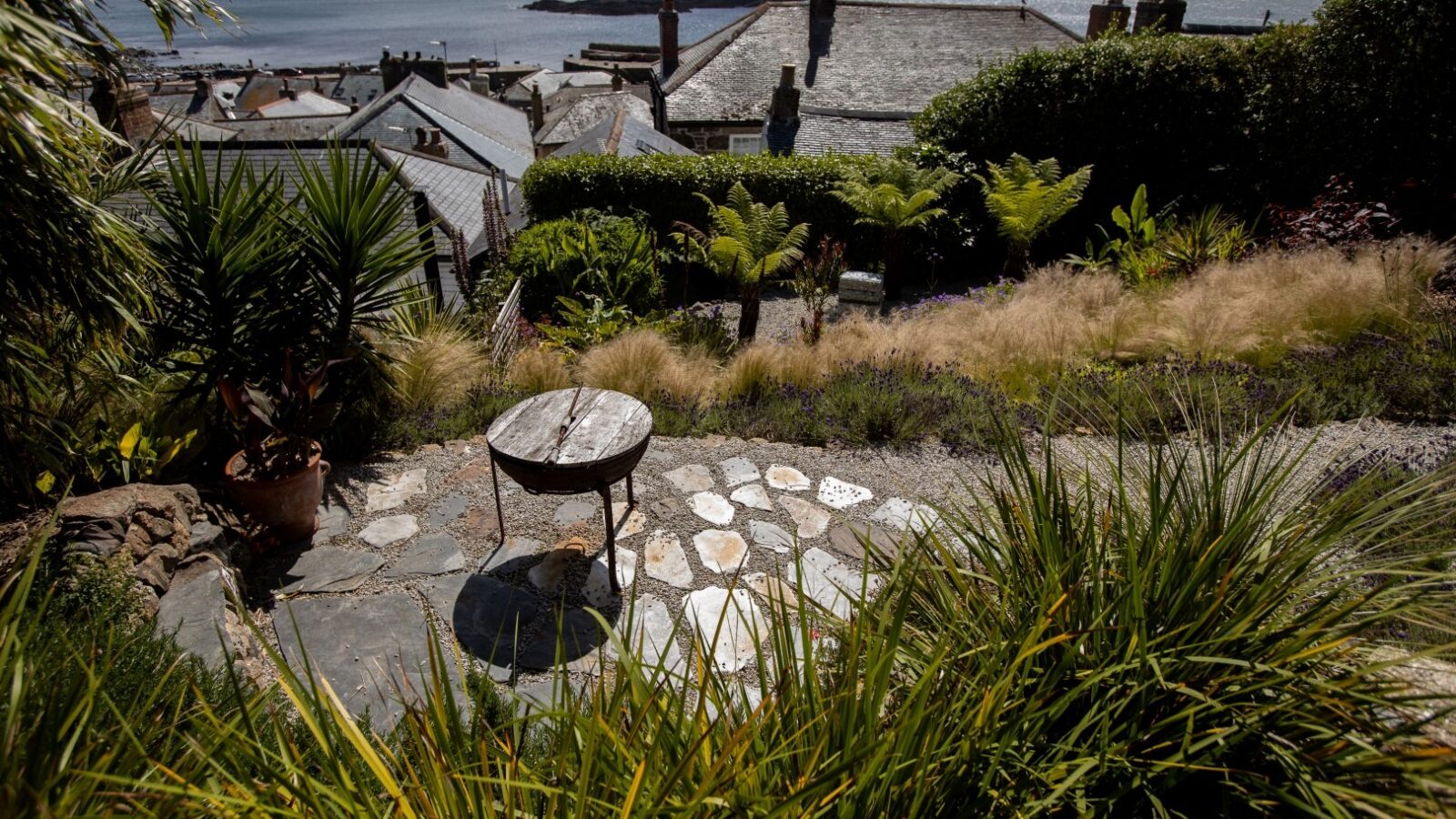 A stone pathway leads to a small circular table in a lush garden. The garden, like a hidden heaven where a fallen angel might find peace, overlooks a tranquil seaside village with quaint rooftops and distant waters under a partly cloudy sky. Greenery, including ferns and palm-like plants, surrounds the pathway.