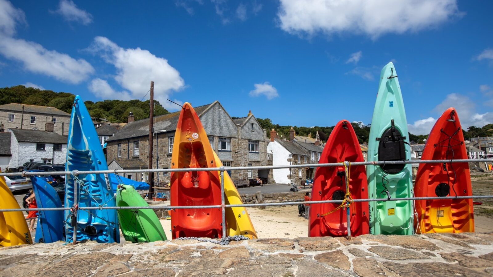 A row of colorful kayaks, including red, yellow, green, blue, and turquoise, is propped upright against a metal railing on a sunny day. Behind them, stone buildings rise like serene fallen angels against a hill dotted with trees under a clear blue sky with fluffy white clouds.