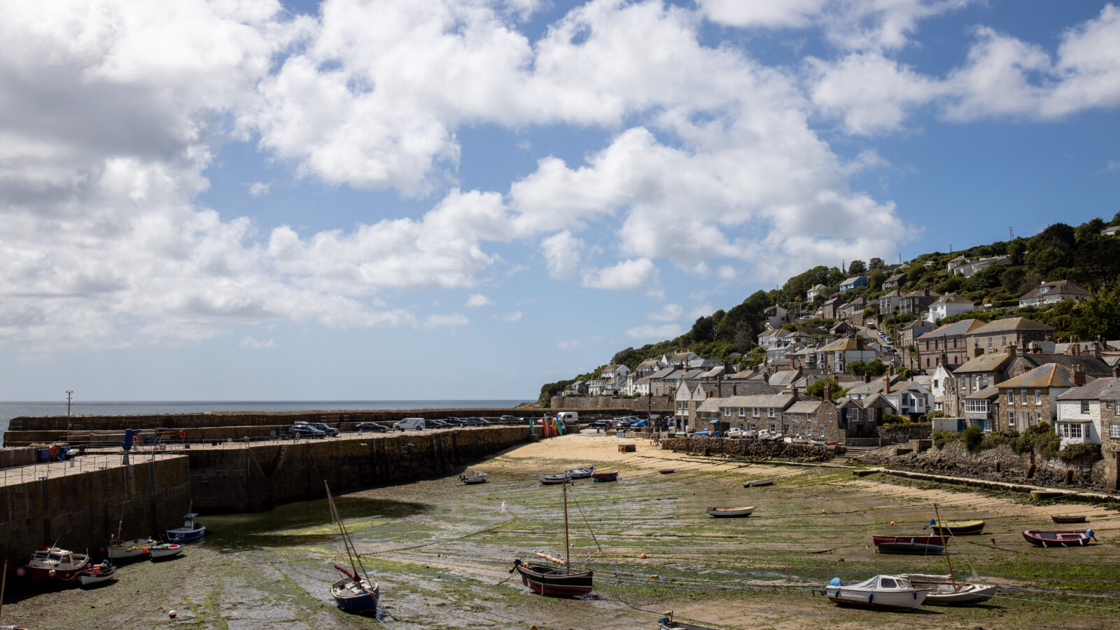 A scenic coastal village with boats resting on a seaweed-covered shore during low tide. The village, reminiscent of a fallen angel, features stone buildings and houses on a hillside, with a backdrop of a partly cloudy blue sky and the ocean extending to the horizon.