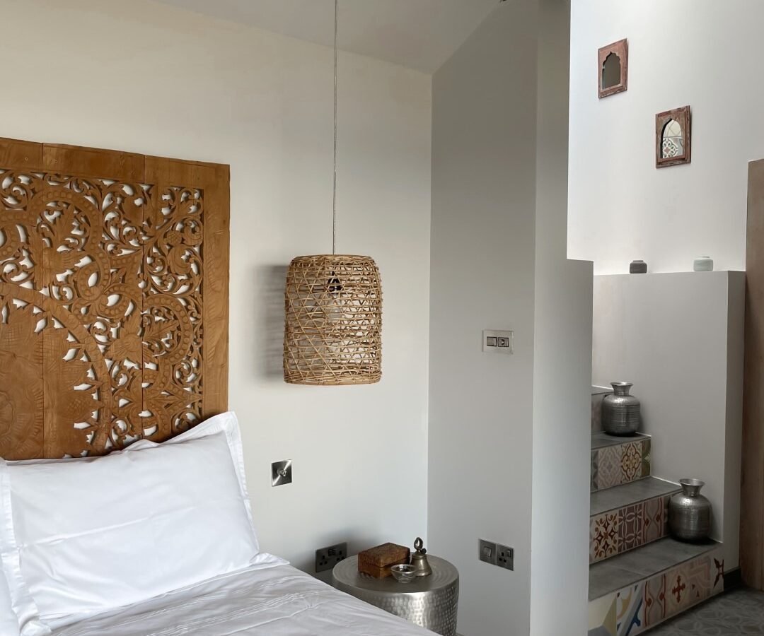 A cozy bedroom featuring a bed adorned with white linens and a carved wooden headboard. A woven pendant light hangs above a small, round metal side table, glowing like an angel. The room has white walls, decorative vases, and patterned tiles on a fallen staircase in the background.