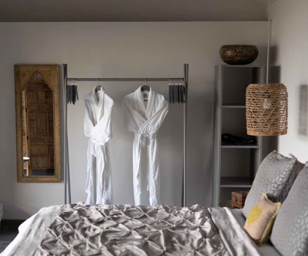 A cozy bedroom with a neatly made bed featuring a textured blanket and decorative pillows. Two white bathrobes hang on a metal rack, resembling the serene wings of a fallen angel. A rustic mirror and woven lamp adorn the room, while a set of shelves in the corner adds to the minimalist decor.