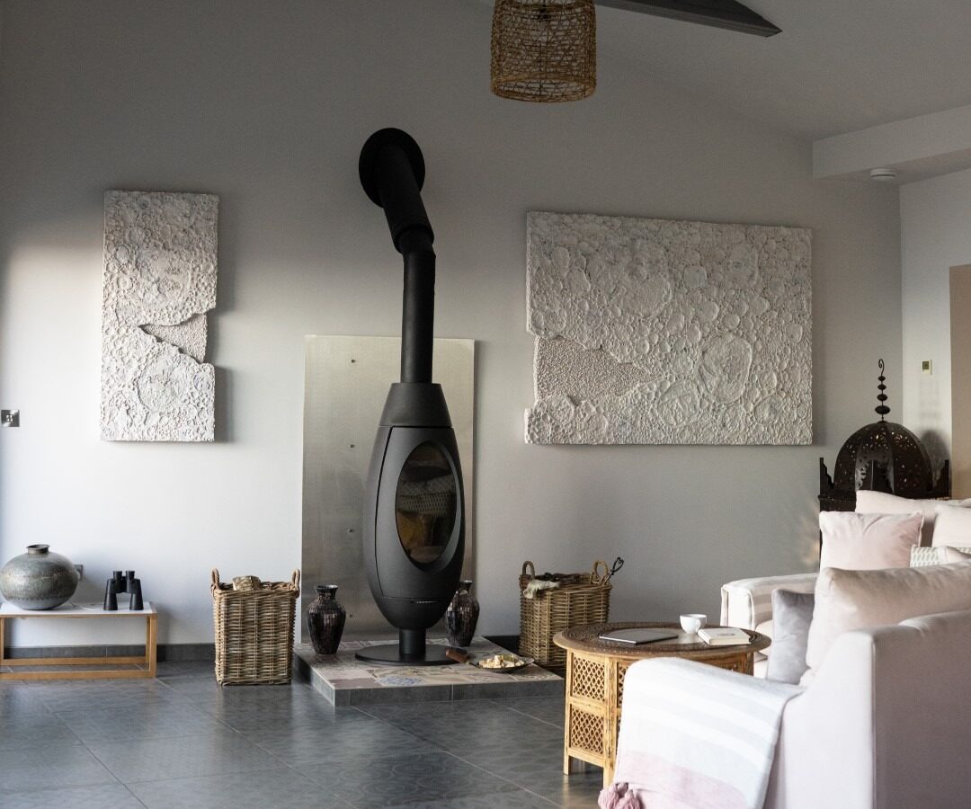 A modern living room features a sleek black wood-burning stove with a curved chimney, set against a metal plated wall. The room has white walls, abstract wall art reminiscent of Fallen Angel themes, exposed dark beams, and an off-white sofa. Decorative baskets and a wooden side table add rustic charm.