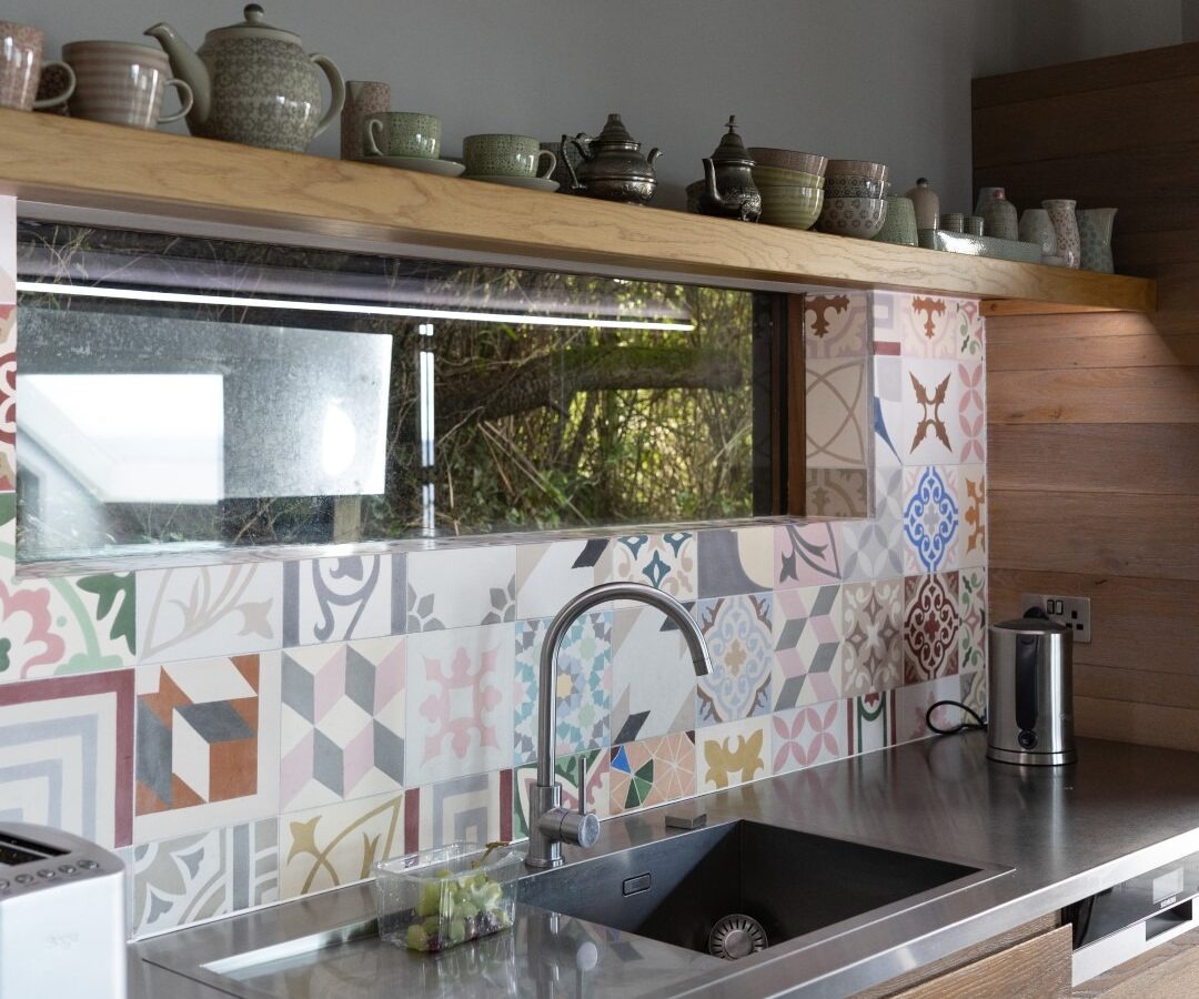 A modern kitchen with wooden cabinets, a stainless steel sink, and a countertop. The backsplash features colorful geometric tiles. Above it, a shelf holds various teapots and cups, while a sculpture of a Fallen Angel hangs on the wall above the shelf.