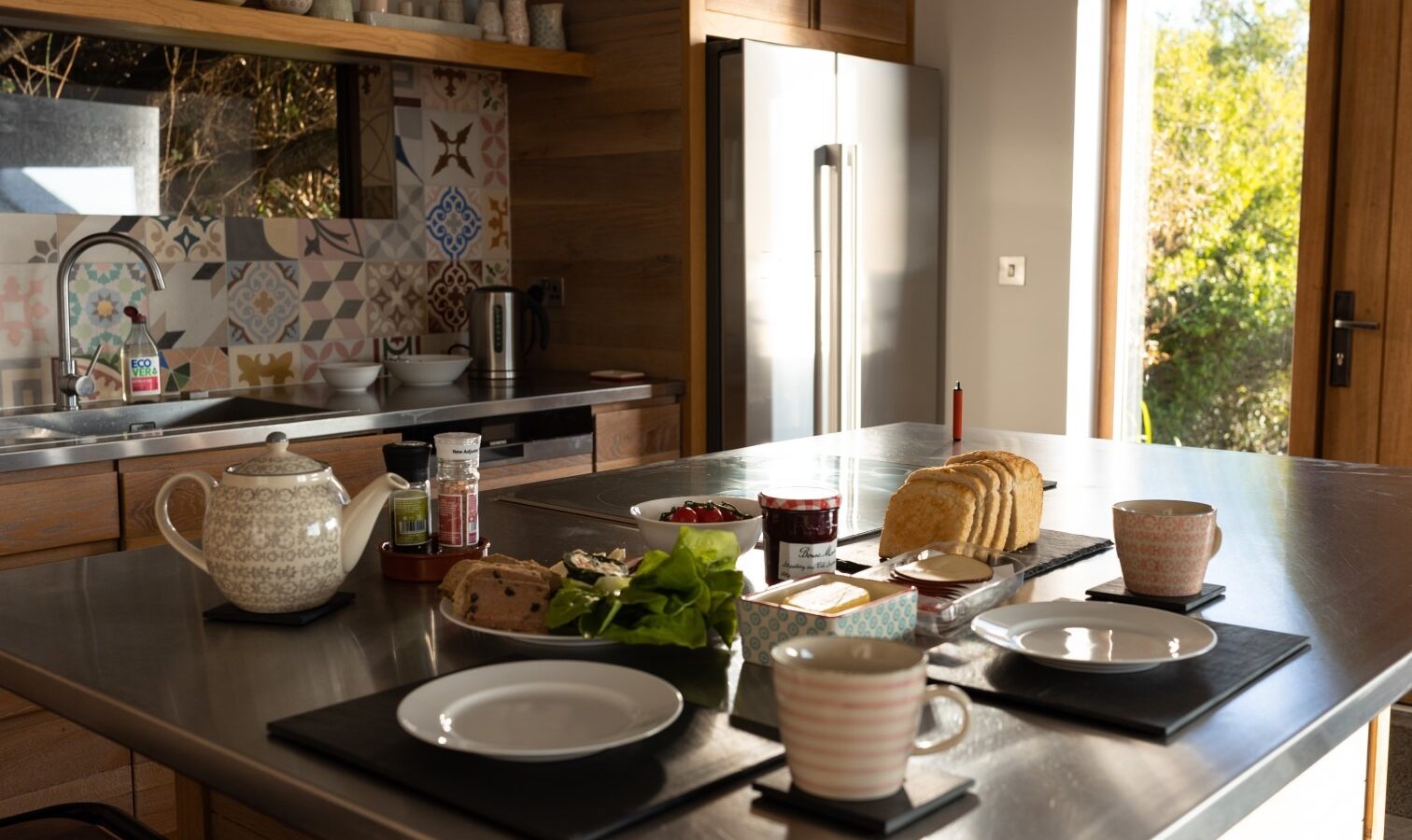 A modern kitchen with a stainless steel countertop set for breakfast. The table includes plates, cups, a teapot, sliced bread on a cutting board, jams, fresh greens, and other condiments. Sunlight streams in from the window like a fallen angel's grace, highlighting the cozy atmosphere.