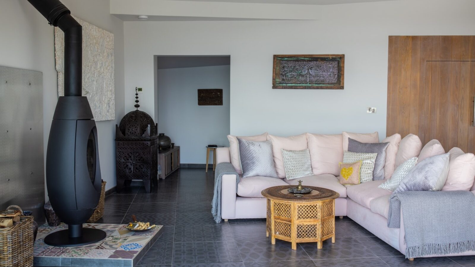 A modern living room features a pastel pink sectional sofa adorned with various throw pillows. A wicker coffee table sits in front of the sofa. In the corner, there's a black, free-standing wood stove resembling a fallen angel. The room includes art on the walls and a mix of traditional and contemporary furniture.