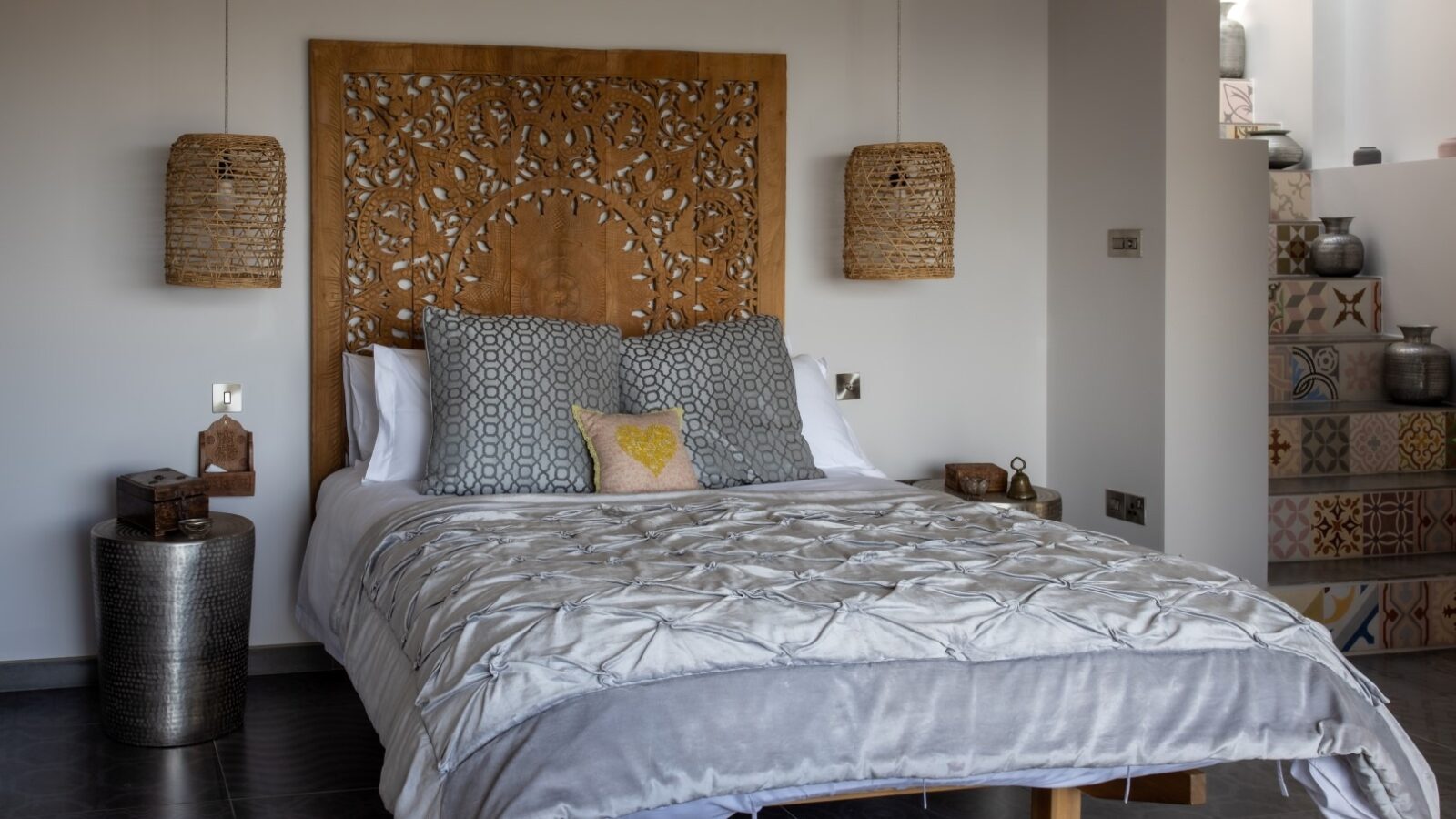 A cozy bedroom featuring a double bed with a patterned wooden headboard and an intricately quilted white comforter that evokes a serene, fallen angel aesthetic. The bed has several pillows in gray and yellow. Two woven pendant lights hang on either side, and decorative items are placed on side tables and steps.