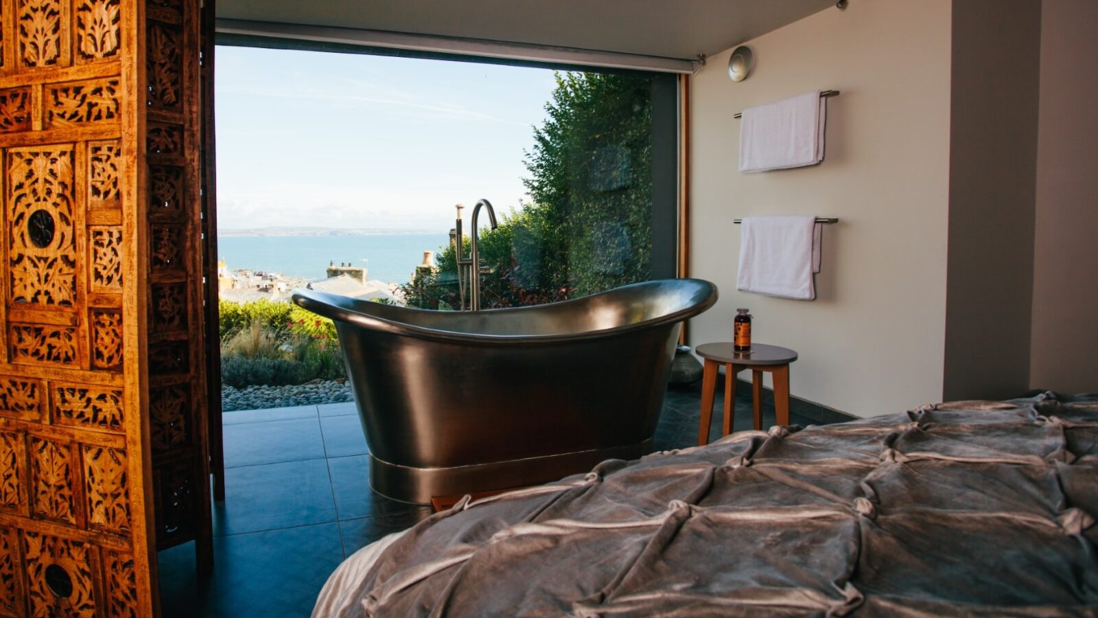 A luxurious bathroom with a freestanding metallic bathtub facing a large window that opens to a scenic coastal view. The foreground features part of a bed with a plush quilt, reminiscent of the comfort found in Fallen Angel’s embrace, and a small wooden table with a bottle placed next to the tub. Two towels hang on the wall.