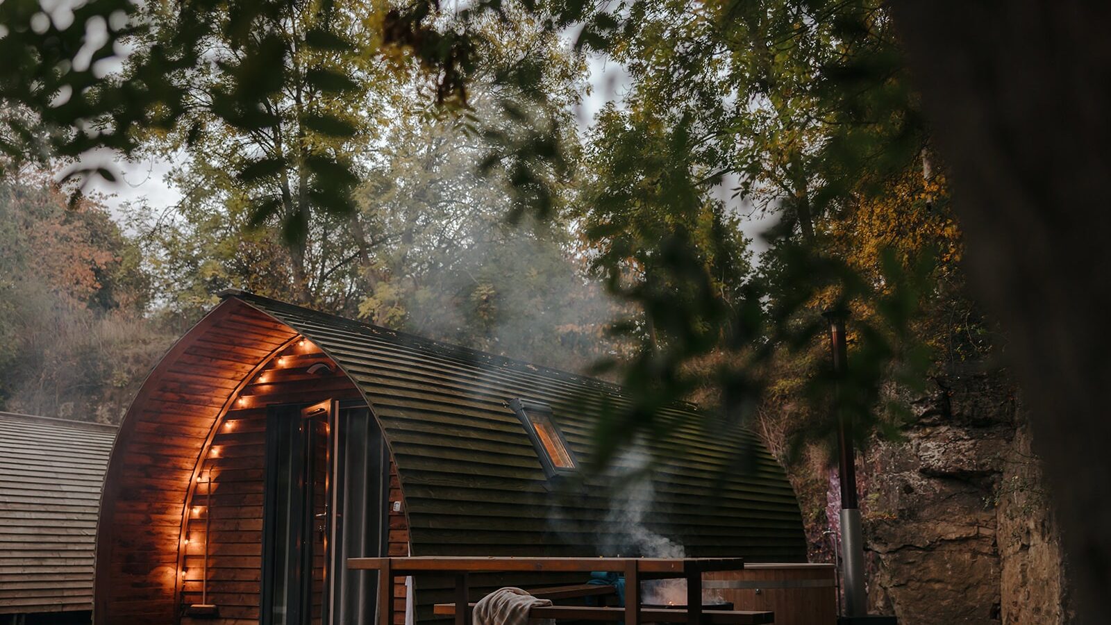 A cozy, wooden, pod-like cabin at Forcett Grange is nestled among trees, with soft exterior lighting illuminating the entrance. Smoke drifts up from a fire pit near a wooden picnic table in the foreground. The setting is serene and inviting, surrounded by autumn foliage.