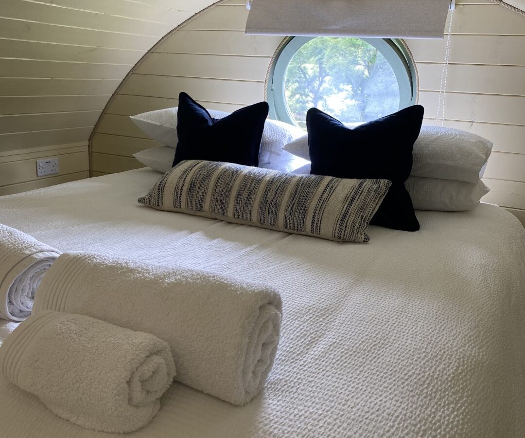 A cozy bedroom with wooden paneled walls, featuring a neatly made bed with white linens, two black accent pillows, and a striped decorative pillow creates an inviting glamping experience. Rolled white towels are placed at the foot of the bed. A round window offers a view of open fields and greenery outside.