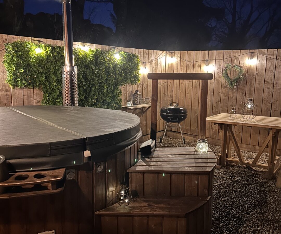 A wooden outdoor hot tub with a closed lid sits in a gravel-covered backyard, perfect for glamping. It has a small set of steps leading up to it. Lit lanterns and string lights create a cozy atmosphere. A green leafy garland decorates the fence, and a small round grill is nearby at Freedom Fields.