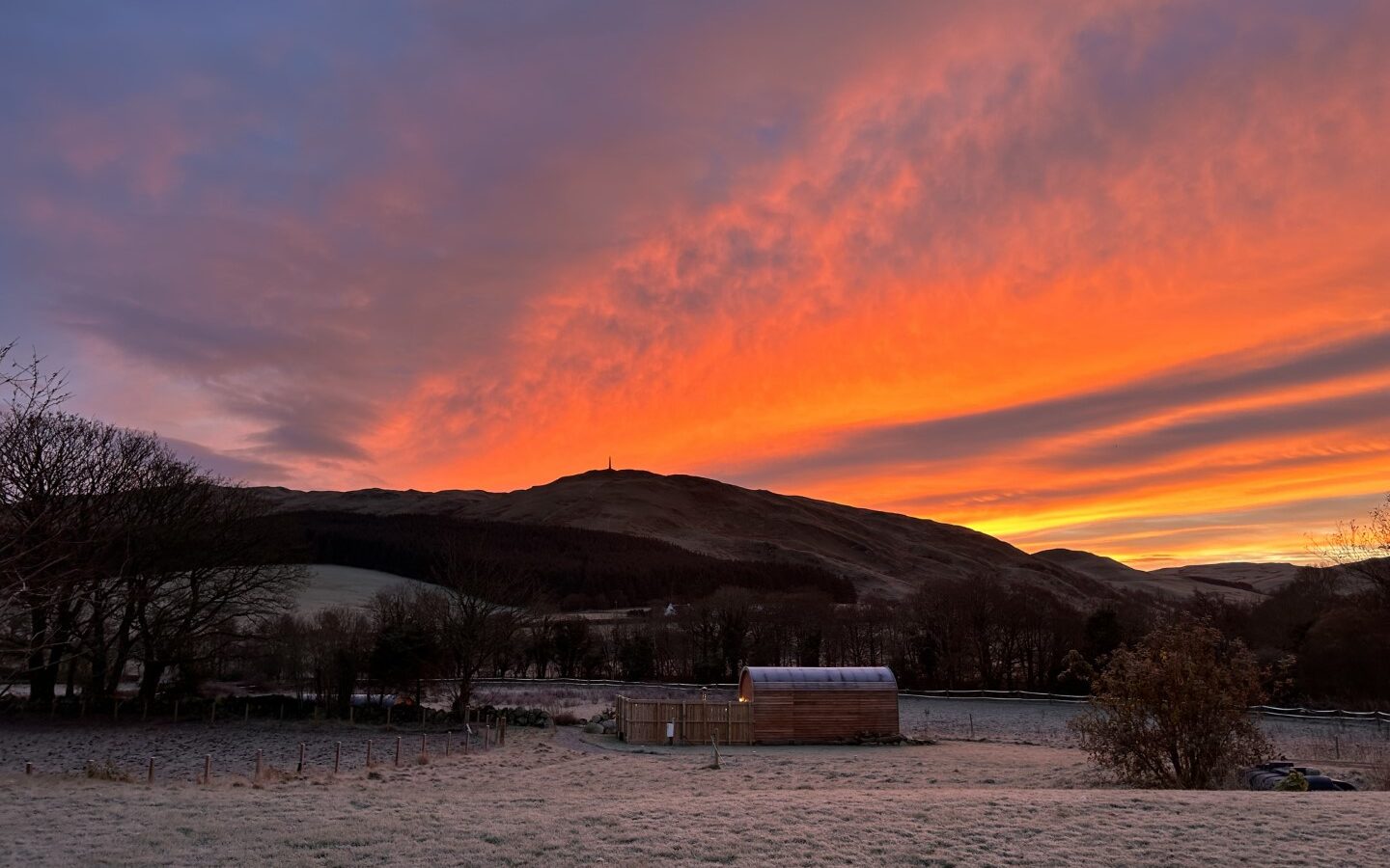 A rural landscape at sunset with vibrant, orange and pink hues in the sky. A small wooden cabin with a greenhouse is in the foreground, part of the charming Freedom Fields glamping site, surrounded by frosty, grassy fields. Hills and leafless trees are visible in the background under the dramatic, colorful sky.