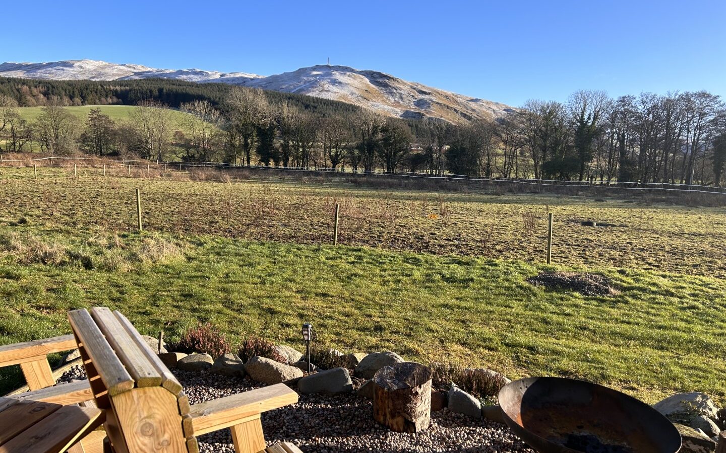 A scenic view of a grassy field with a snow-capped mountain in the background under a clear blue sky at Freedom Fields Glamping. In the foreground, there's a wooden bench next to a fire pit. Trees are scattered throughout the landscape.