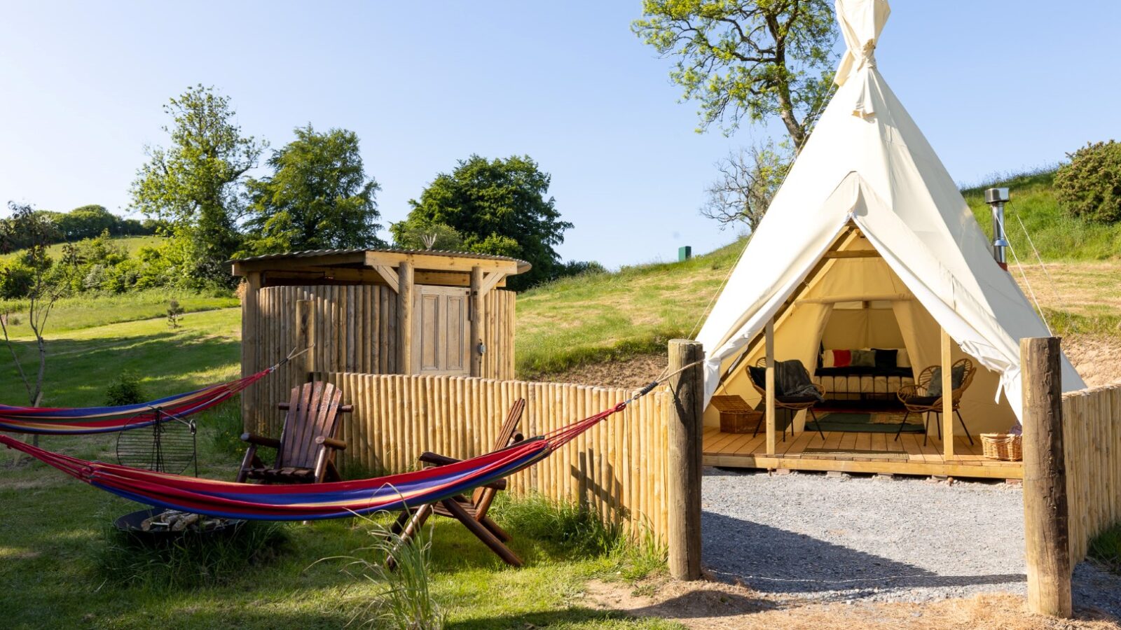 A serene HARTA retreat campsite featuring a white, canvas tipi with open flaps reveals a cozy interior. Two colorful hammocks are stretched between wooden posts near a rustic shed. Surrounding lush greenery and a clear blue sky complete the tranquil scene.