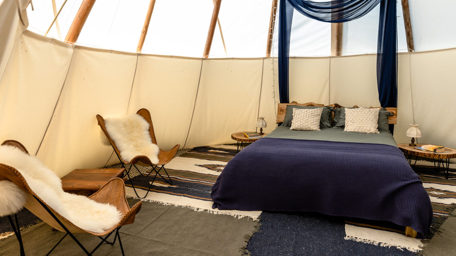 Experience a cozy retreat inside an HARTA tent with a wooden frame. The bed, adorned with a blue blanket, gray pillows, and a wooden headboard, promises comfort. Blue drapes hang above the bed. The room features two brown butterfly chairs with white fur throws and two small wooden side tables.