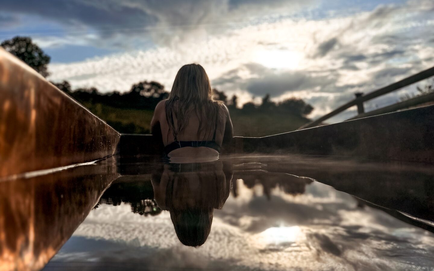 A person with long hair, seen from behind, sits in a water slide at Hadspen. The image captures the reflections of the sky and trees in the water, with clouds and lush forest in the background. The evening sky is filled with soft, dramatic clouds and sunlight breaking through, adding a touch of glamping tranquility.