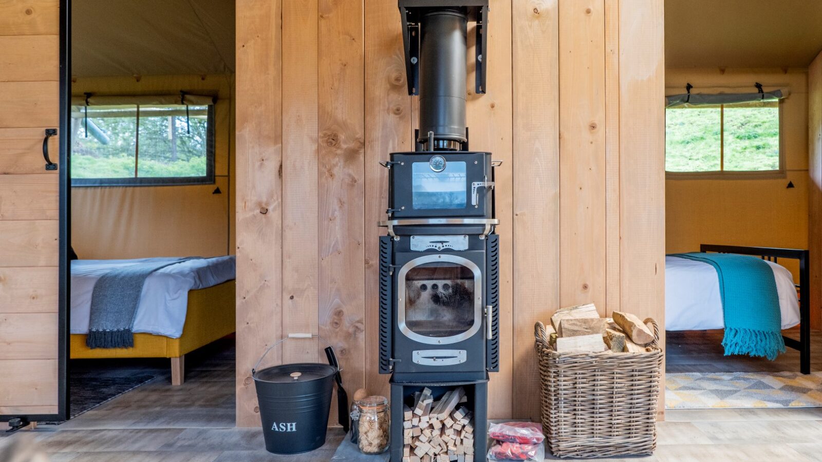 A cozy Hadspen Glamping interior features a wood-burning stove centered against a wooden wall. Firewood is stored below the stove, with a basket of logs and an ash bucket on either side. Two adjacent rooms are visible, each with a neatly made bed by the windows.