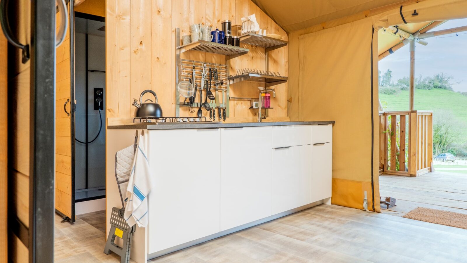 A cozy kitchen in a wooden cabin tent at Hadspen. It features white cabinets, a countertop with a kettle and various dishes, and utensils hanging on a rack. The tent opens to a porch with a scenic outdoor view, perfect for glamping. The flooring is light-colored and the walls are wooden.