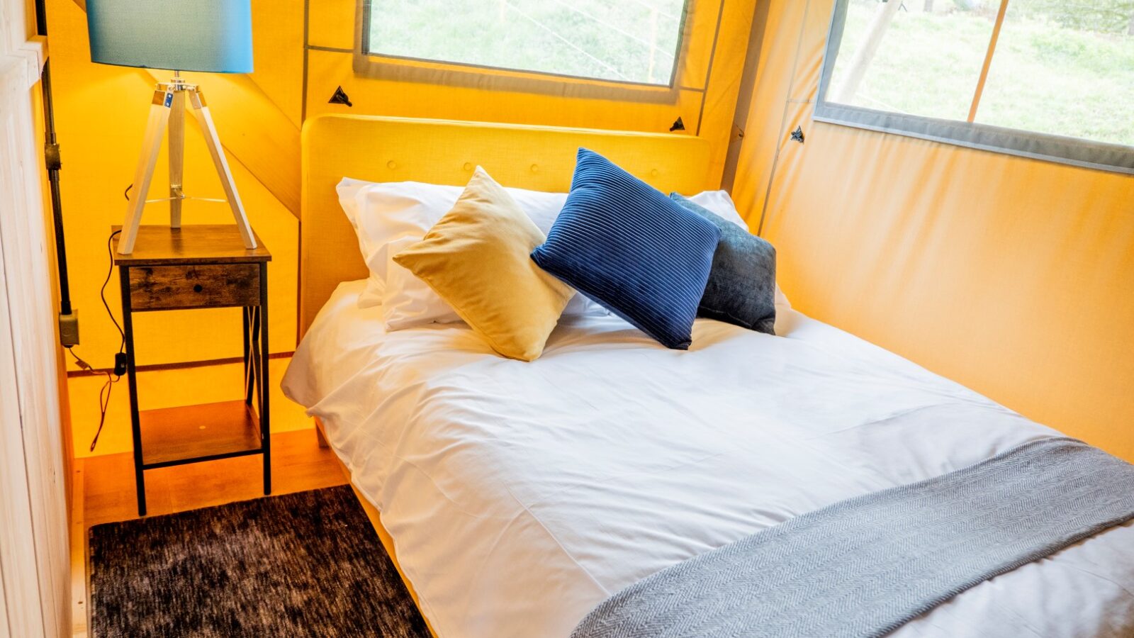 A cozy bed with white linens and a gray blanket is adorned with yellow, blue, and gray decorative pillows. A modern lamp with a blue shade sits on a wooden side table beside the bed. The glamping tent at Hadspen features large windows in its yellow walls, creating a bright and airy atmosphere.