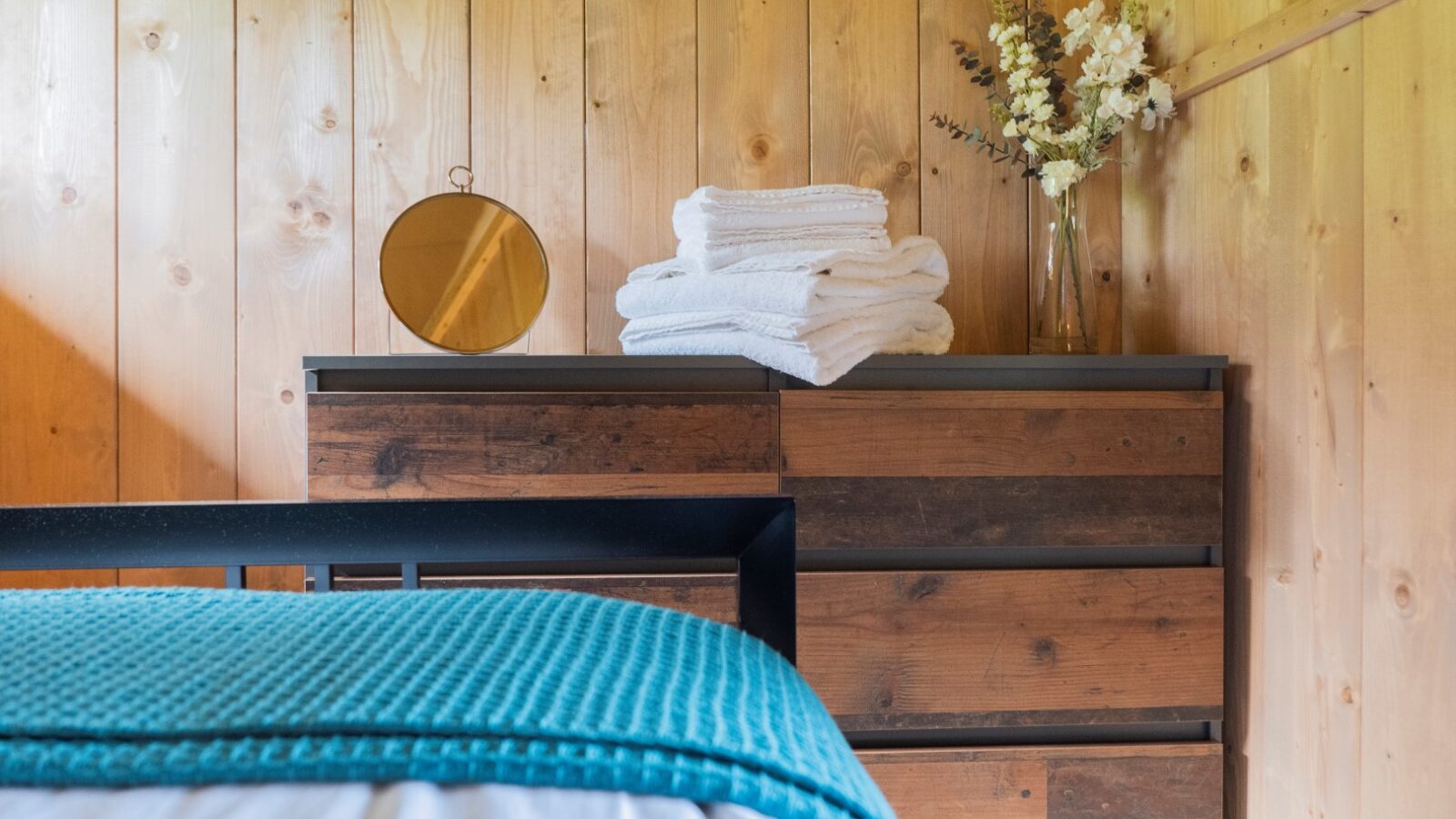 A cozy bedroom corner in Hadspen features a wooden dresser with a round mirror, neatly folded white towels, and a vase of white and beige flowers. The bed's blue blanket and white sheets are partially visible in the foreground, enhancing the glamping experience against light wood panel walls.