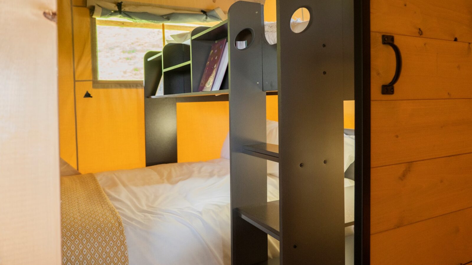 This image shows a cozy interior of a small glamping cabin or tent in Hadspen, with wooden walls and a bed made up with a white sheet and a patterned duvet cover. Adjacent to the bed is a dark-colored storage unit featuring shelves and cubby holes, partially obstructing the bed.