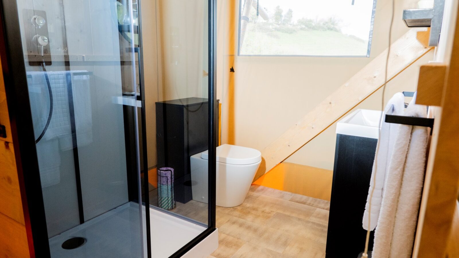 A modern, compact bathroom at Hadspen Glamping features a glass shower enclosure on the left, a toilet in the center beside a slanted wooden ceiling beam, and a sink with a white towel on the right. Natural light pours in through a window with views of lush greenery outside.