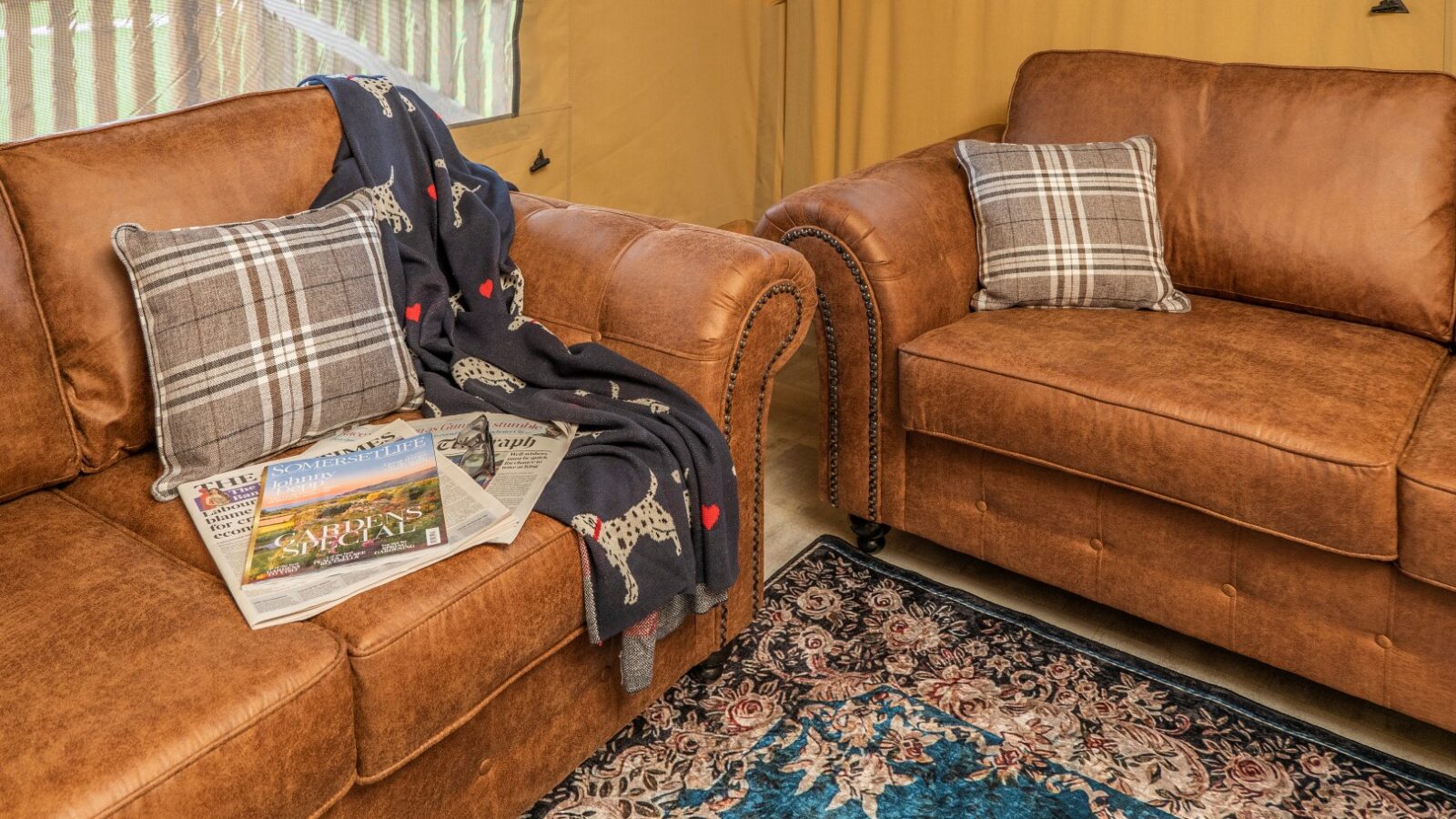 Two brown leather sofas adorned with plaid throw pillows create a glamping vibe in this cozy Hadspen room. One sofa has a black blanket with dog patterns and a stack of newspapers and magazines. A patterned rug covers the floor, and large windows offer views of the lush green outdoor area.