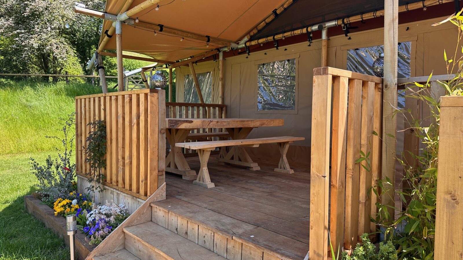 A cozy outdoor wooden deck with steps leads to a shaded seating area under a canvas canopy. The deck features a wooden table and benches. Surrounding the deck are vibrant flowers, lush greenery, and a well-maintained lawn, providing a serene and inviting atmosphere—perfect for your Hadspen Glamping experience.