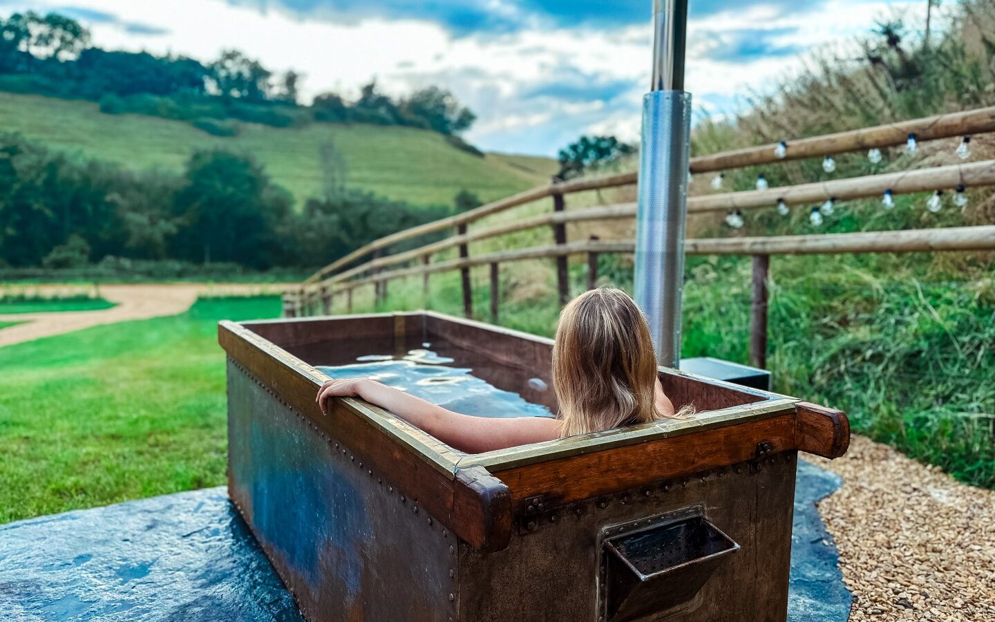 A person with blonde hair is soaking in a large, rustic outdoor bathtub with a metal chimney at a charming Hadspen glamping site. The tub is nestled in a scenic countryside setting with rolling green hills and a partly cloudy sky. A wooden fence lines the path beside the tub.