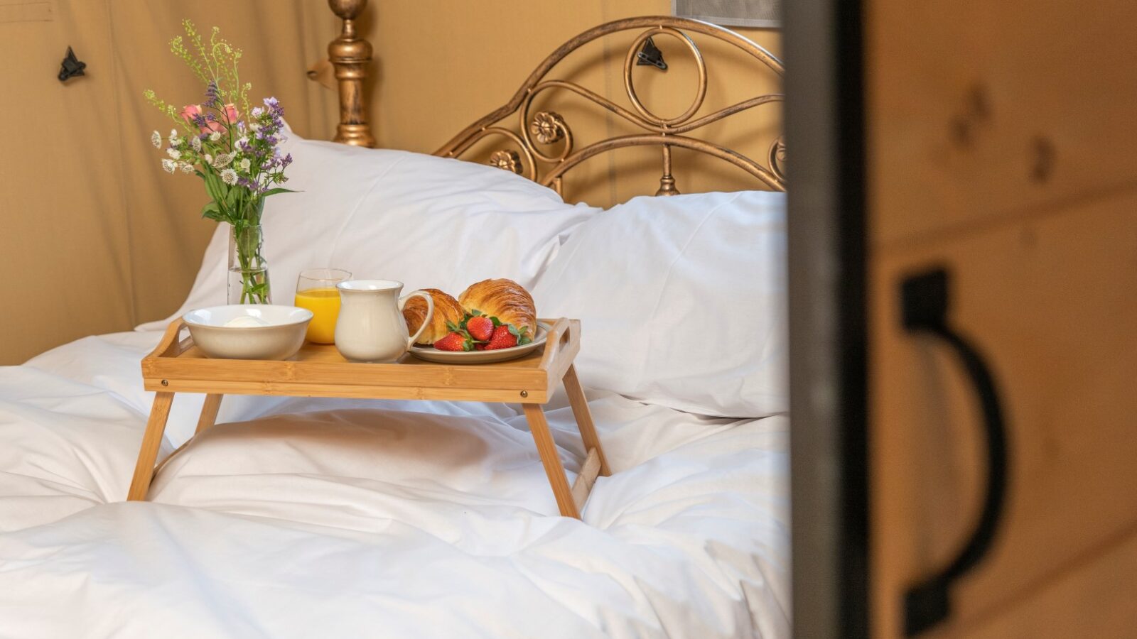 A cozy bed with white linens features a wooden breakfast tray holding croissants, strawberries, a bowl, a pitcher, and a glass of orange juice. A small vase with flowers is also placed on the tray. The room has warm-toned decor and a classic metal headboard, embodying the charm of Hadspen Glamping.