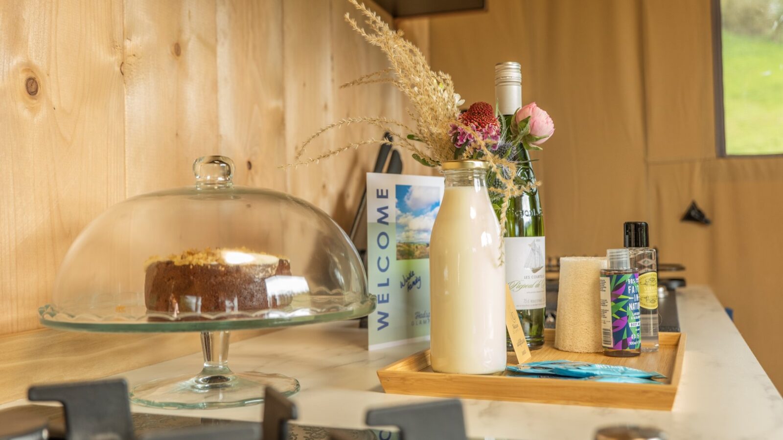 A cozy kitchen table in a rustic Hadspen setting holds a glass cake stand with a cake, a glass bottle of milk, a small bouquet of flowers in a vase, wine bottles, a welcome brochure, and a tray with salt and pepper shakers. The background features wooden paneling and hints at glamping comfort through the window.