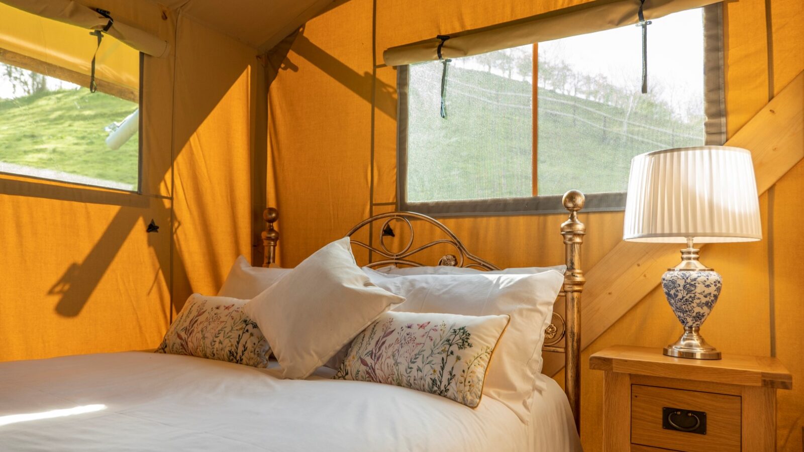 A cozy, warmly-lit tent interior in Hadspen features a metal-framed bed with white linens and floral throw pillows. Beside the bed is a wooden nightstand with a blue and white patterned lamp. Sunlight filters through the tent's windows, revealing a grassy hillside outside—glamping at its finest.