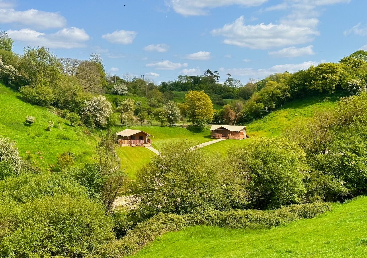 Scenic landscape showing two small wooden cabins nestled in a lush, green valley near Hadspen. The area is surrounded by rolling hills, dense greenery, and trees. The sky is blue with scattered white clouds, suggesting a sunny, pleasant day perfect for glamping in nature.