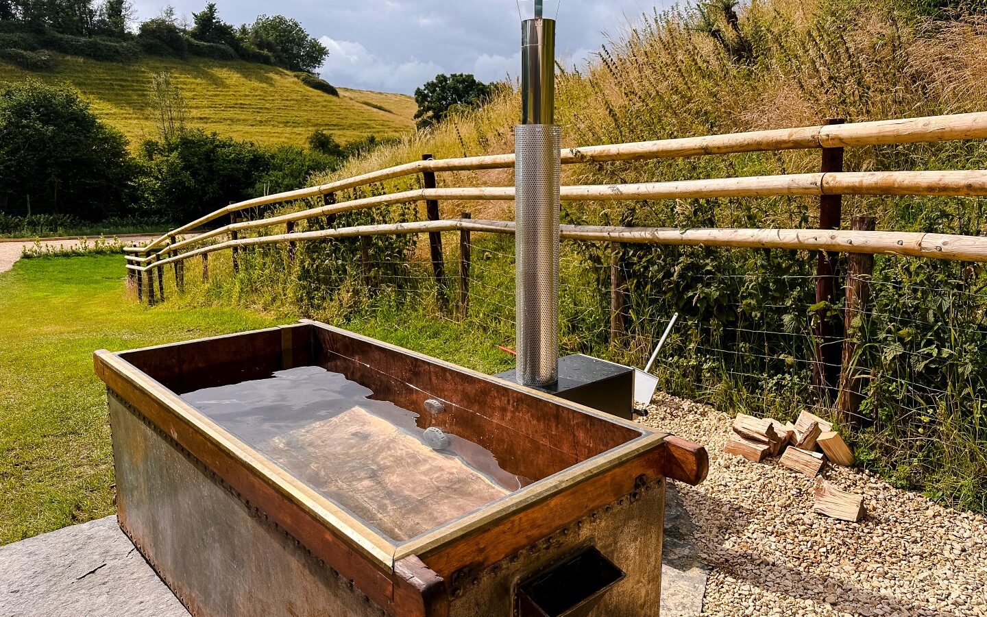 An outdoor wooden hot tub with a metal stove and chimney is set on a gravel area beside a wooden fence. The grassy hills and cloudy sky in the background create a serene countryside atmosphere, perfect for glamping near Hadspen. Logs of wood are stacked beside the tub, ready for use.