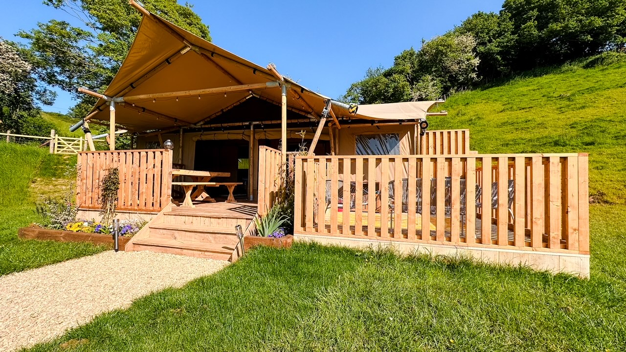 A glamping tent on a lush green hillside in Hadspen, featuring a wooden deck with railings, outdoor seating, and potted plants. The tent has an open front with a picnic table on the deck and a canopy overhead, blending rustic wood and canvas elements. Trees and grass surround the area.