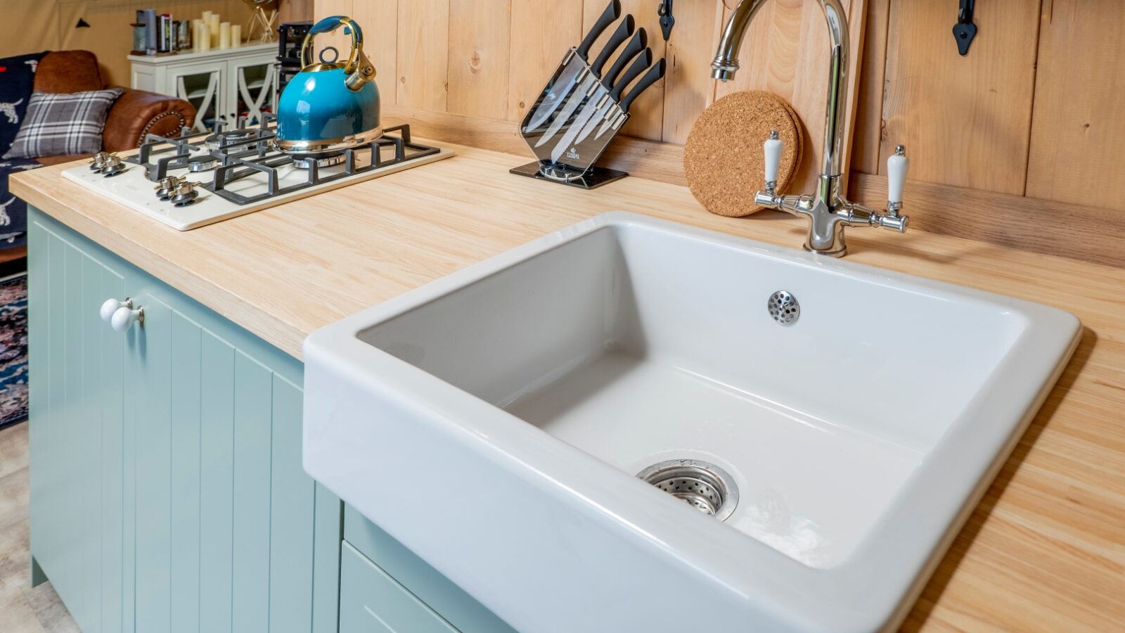 A modern kitchen counter in Hadspen features a large white farmhouse sink, a sleek chrome faucet, a set of knives in a wooden block, and a teal kettle on a gas stove. The countertop has a light wood finish, and the cabinets are painted pastel green, creating the perfect glamping atmosphere.