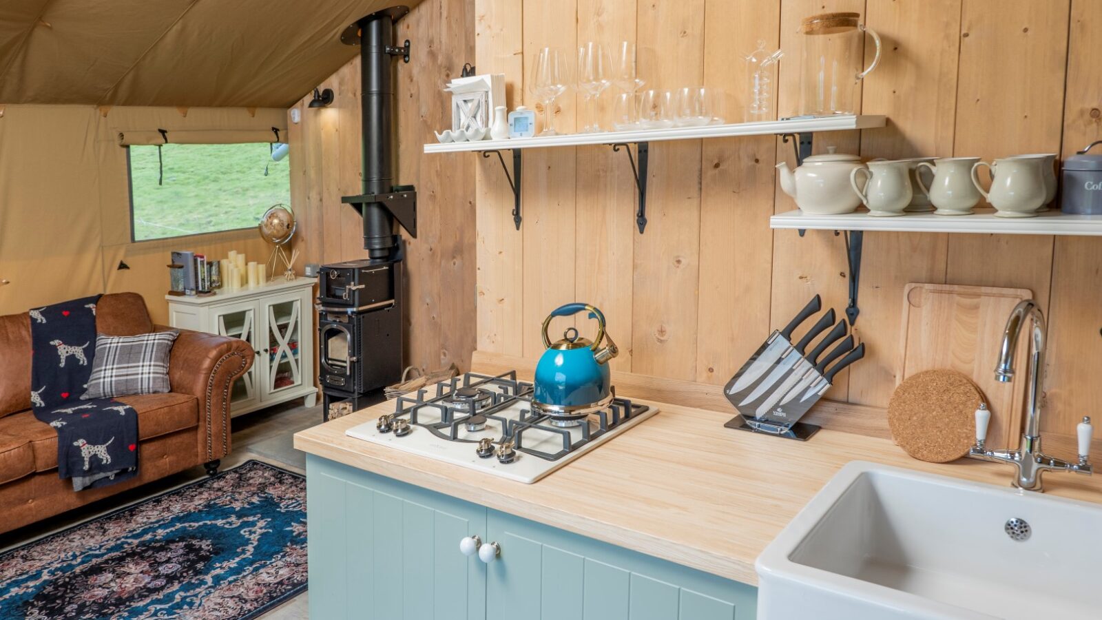 A cozy glamping kitchen and living area inside a tent at Hadspen features a light blue cabinet with a farmhouse sink, a stovetop with a blue kettle, and shelves holding glasses and mugs. The living area has a brown leather sofa, a wood-burning stove, and a rug with a blue pattern.