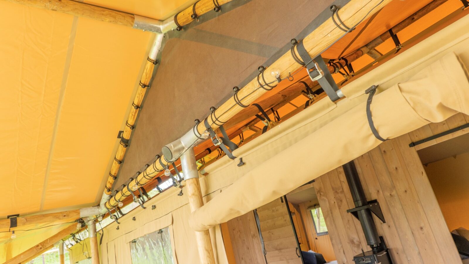 The image shows the interior of a Hadspen glamping tent with a light wooden and canvas structure. The open space features a cozy bed, a wood-burning stove, and a small seating area. The tent's frame is visible, with numerous metal brackets and electrical wiring against the canvas roof.