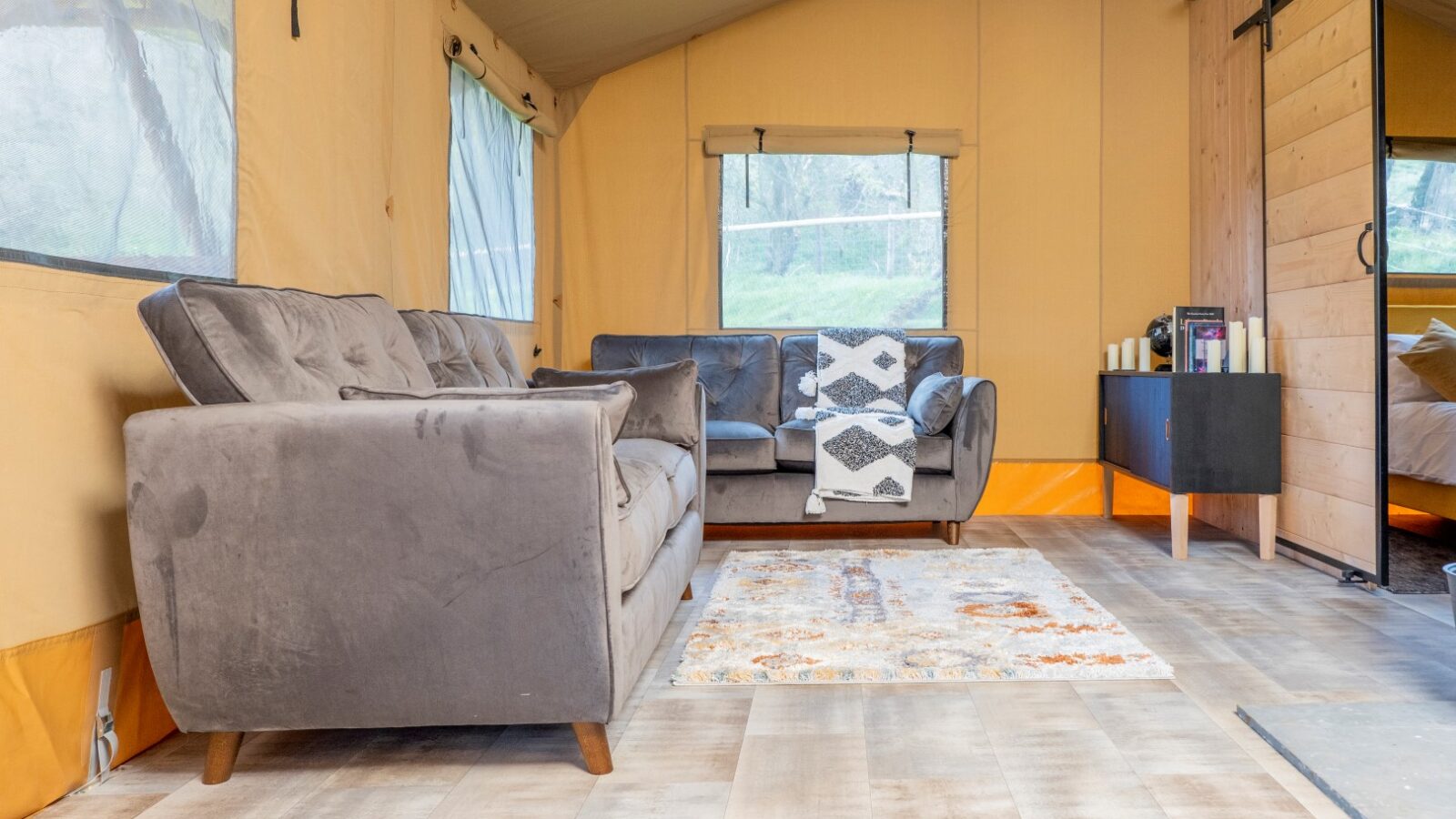 A cozy tent interior in Hadspen features two grey sofas, a colorful area rug, and a wooden side table with candles. Light filters through large windows, illuminating the space. The wooden floor and soft furnishings create a warm, inviting atmosphere perfect for glamping enthusiasts.