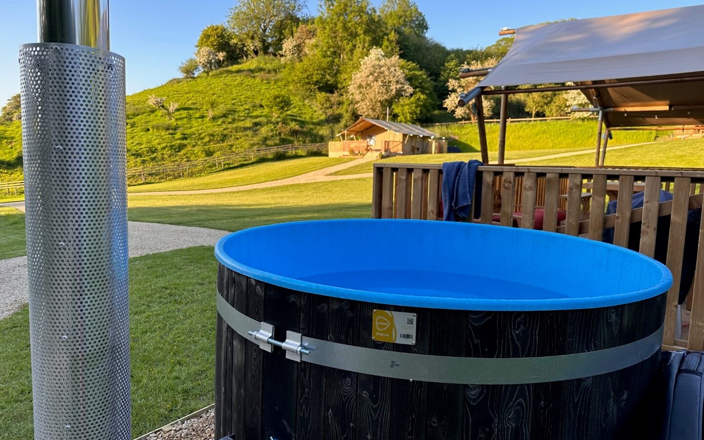 A blue circular hot tub sits on a grassy lawn next to a cylindrical metal structure, possibly a heater. In the background, there is a wooden structure with a canopy perfect for glamping, and an open field with trees and hillsides under the clear blue sky typical of Hadspen.