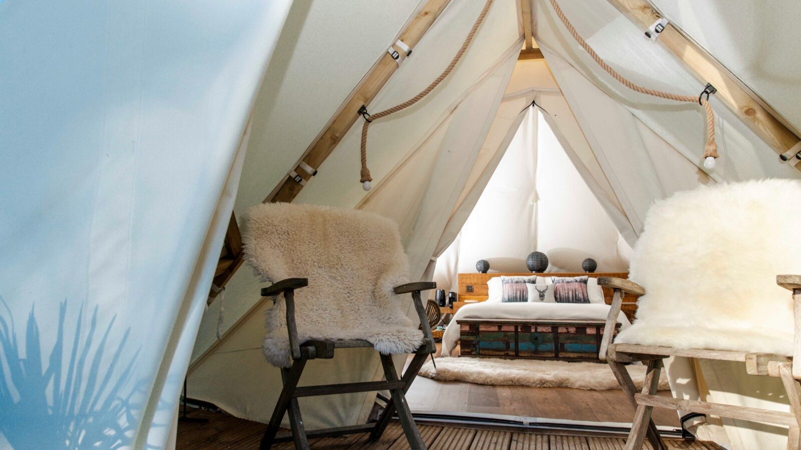 Inside view of a HARTA glamping tent with wooden chairs covered in fluffy cushions, set on a wooden deck. The tent reveals a cozy interior with a double bed, pillows, and soft lighting, giving a warm and inviting atmosphere. Shadows of plants are visible on the tent fabric, perfect for a retreat.