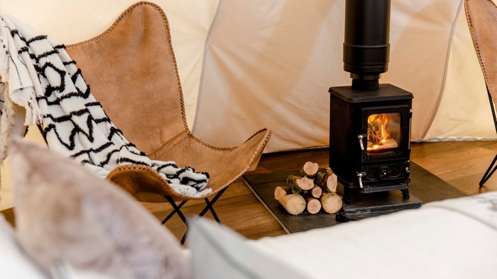 A cozy tent interior with a lit wood-burning stove and a pile of firewood beside it. A leather butterfly chair with a black and white patterned blanket draped over it sits nearby, creating a warm and inviting retreat. Soft cushions from HARTA are partially visible in the foreground.