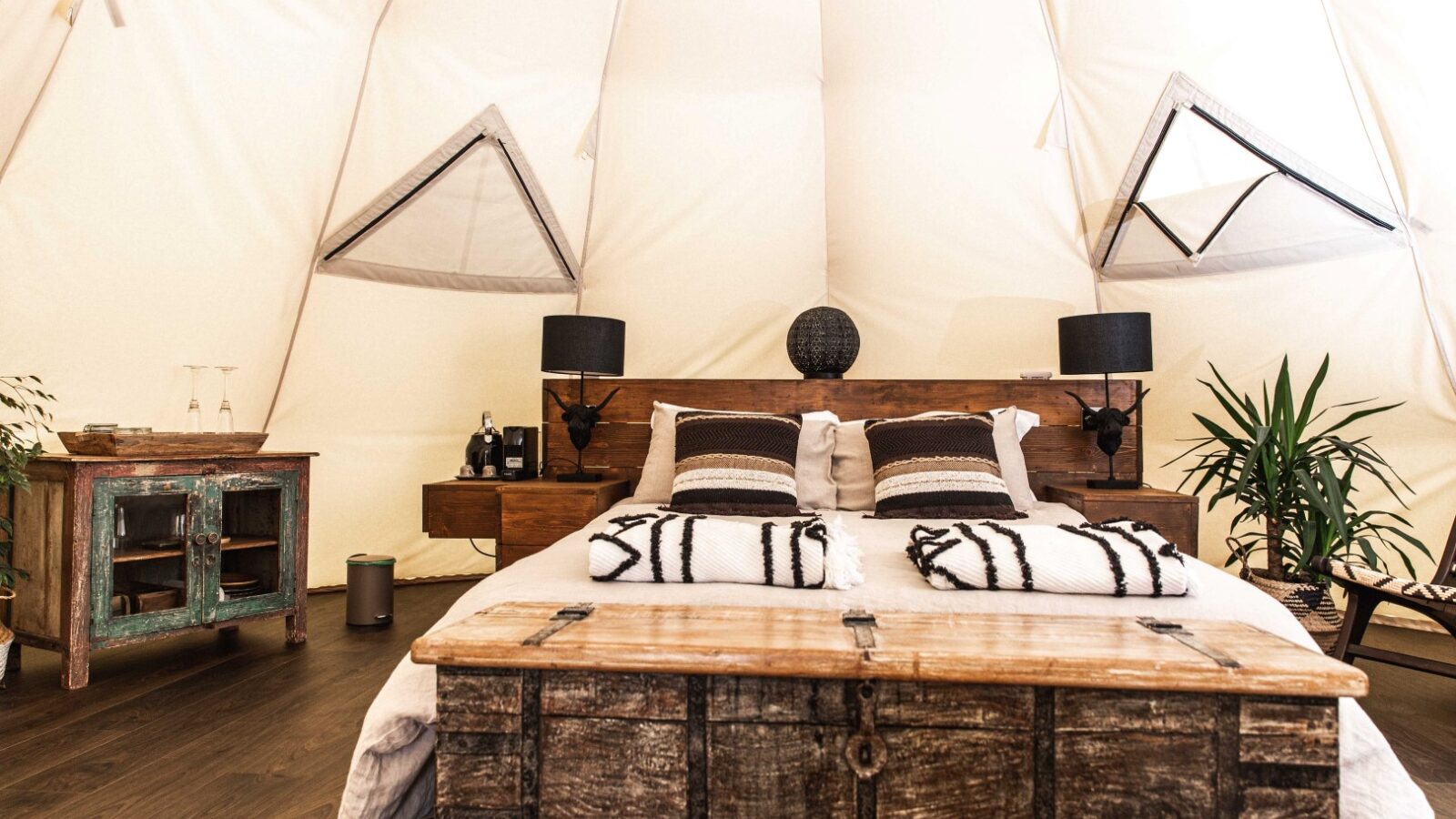 A cozy glamping tent with a neatly made bed featuring black and white pillows and blankets. The room has rustic wooden furniture, including bedside tables with black lamps, a chest at the foot of the bed, and a small cabinet. Sparse greenery adds a touch of nature to this serene HARTA retreat.