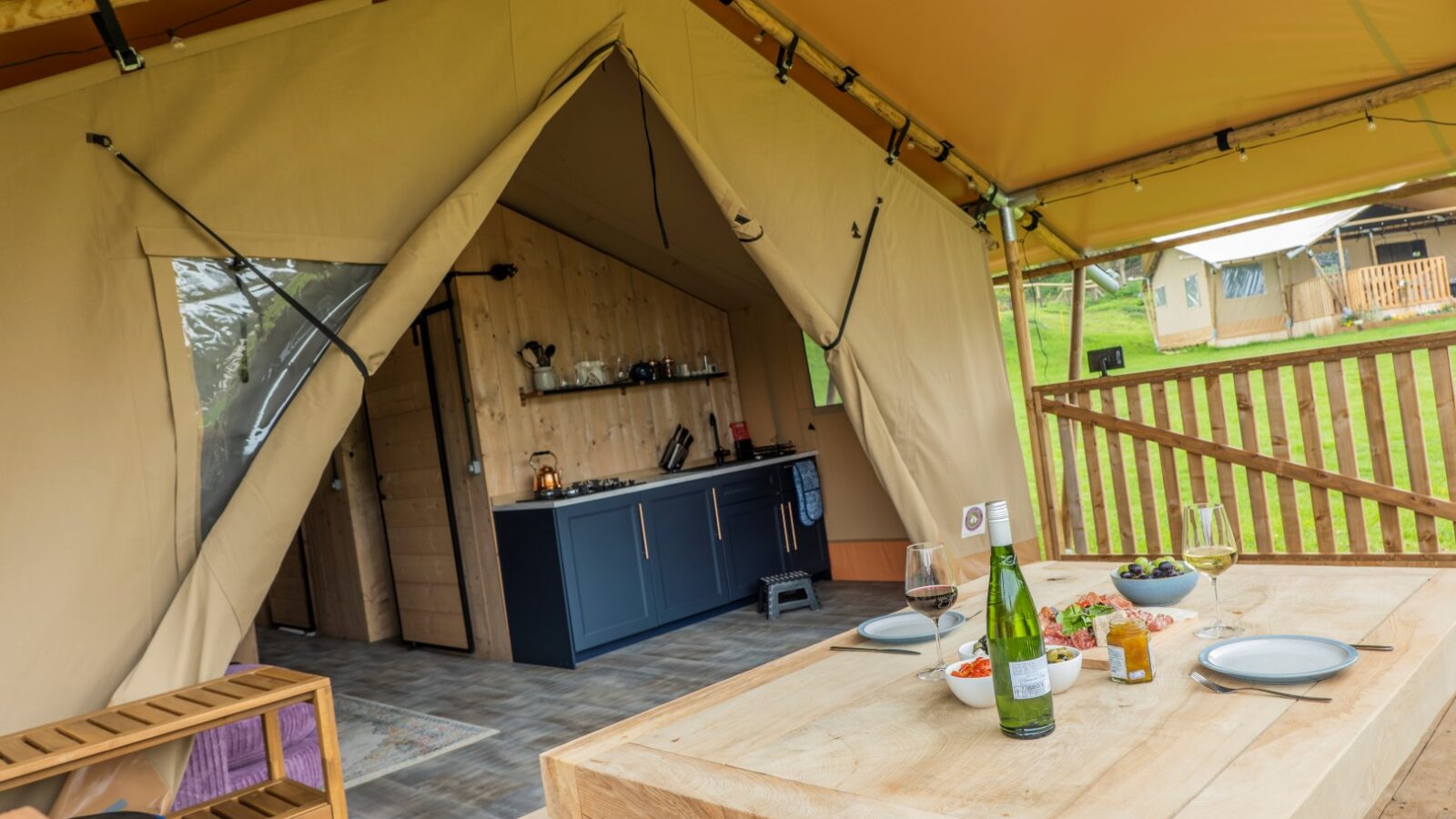 A glamping tent in Hadspen with an open front reveals a rustic interior. The kitchen area with shelves, a stove, and utensils is visible. A wooden dining table is set with plates, glasses, and a wine bottle. The tent is pitched on a grassy area with another tent in the background.