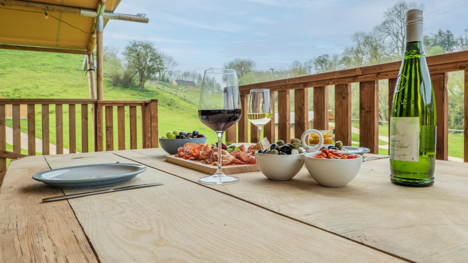 A wooden table is set with two glasses of wine, a bottle of wine, plates, and bowls of olives, tomatoes, and charcuterie. The glamping setup is on a balcony overlooking Hadspen’s green fields with trees under a yellow canopy. The atmosphere is serene and inviting.