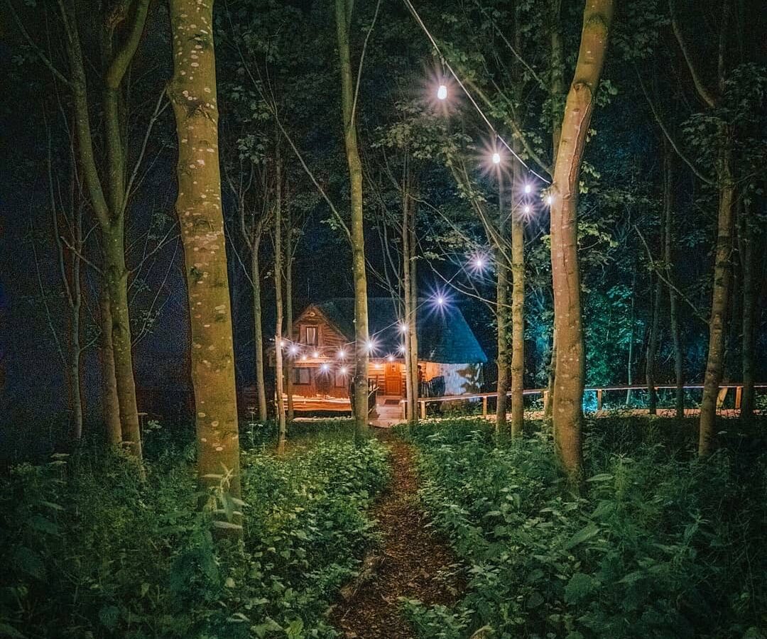 A narrow dirt path leads through a dense forest at night towards a dimly lit house in the distance. Strings of hanging lights illuminate the trees, creating a magical atmosphere. The scene is serene and inviting, almost as if it were leading to Rufus's Roost or Baxby Manor.